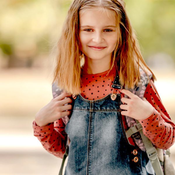 Что подарить на День Рождения девочке - симпатичная милая светловолосая девочка в джинсовом сарафане с красной майкой и длинными рукавами, улыбается оборачиваясь с рюкзаком на фоне парка
