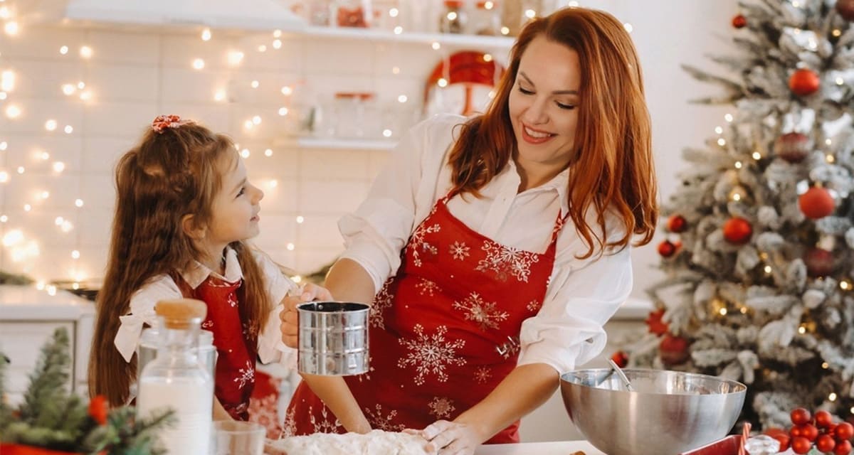 Новогодние подарки маме - мама с дочной готовят новогоднее тесто в нарядно украшенной кухне рядом с елкой