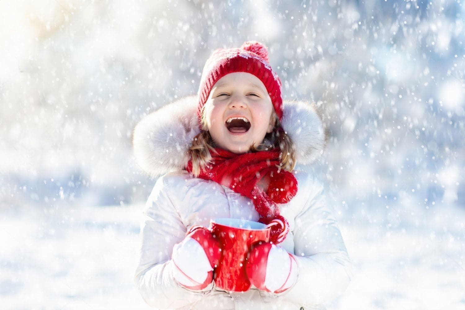 Что подарить девочке на новый год - радостная девочка в белом с красной шапкой и шарфом под снегопадом на солнце держит красную чашку